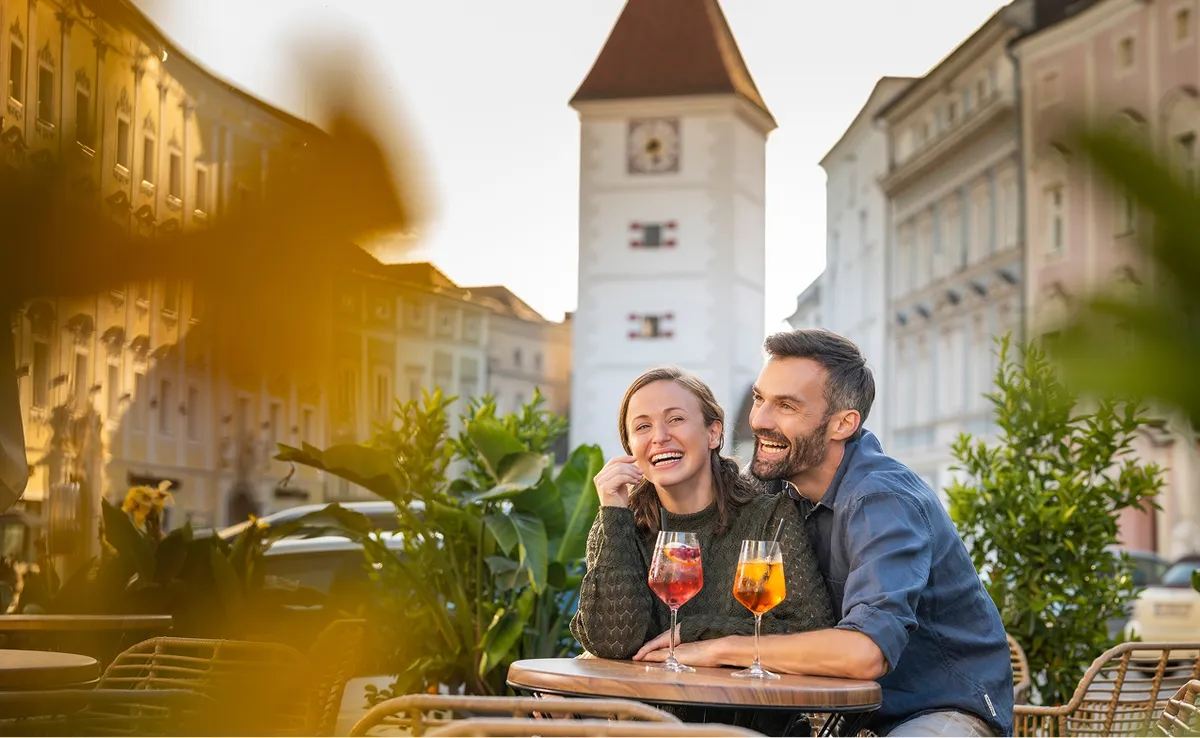 Zwei glückliche Menschen genießen im sonnigen Gastgarten Wels’ charmante Altstadt Drinks und gemütliche Atmosphäre.