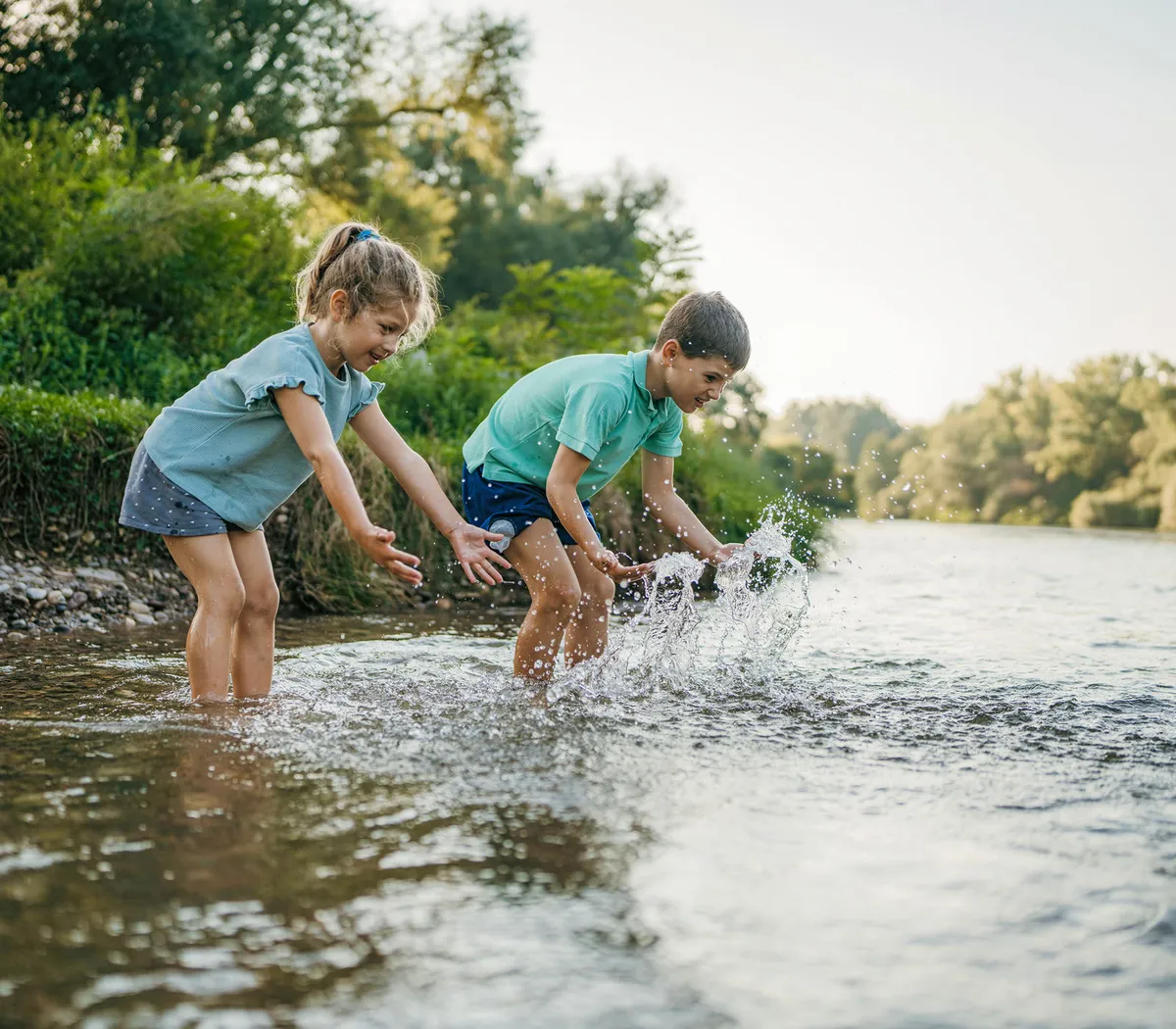 Zwei Kinder spielen lachend im flachen Wasser eines klaren Flusses, umgeben von Natur und Grün.