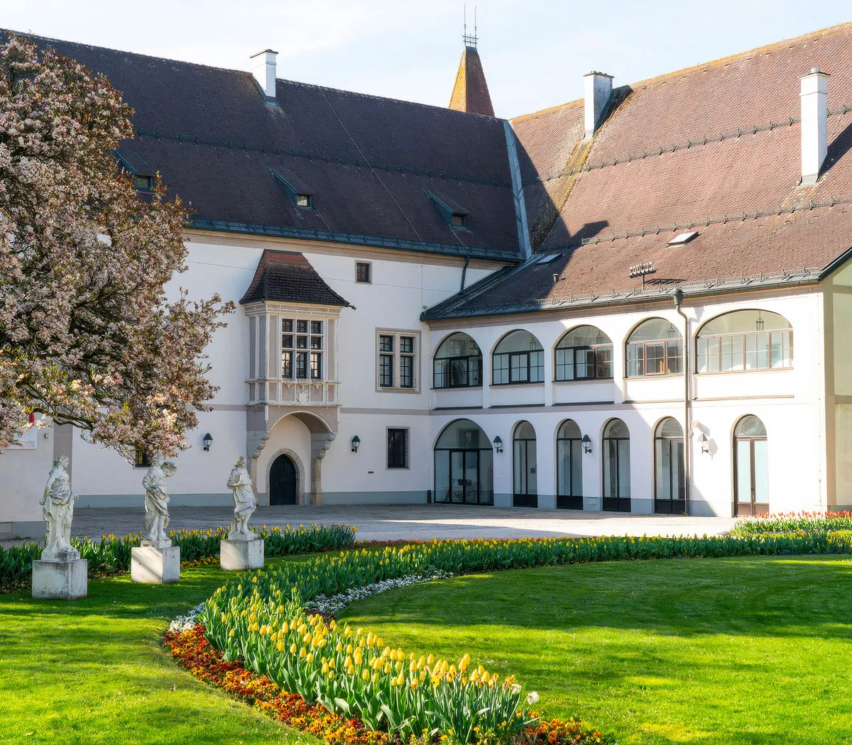 Das Bild zeigt den gepflegten Burggarten Wels während der Landesgartenschau mit Blumenbeeten, Skulpturen und historischem Gebäude.
