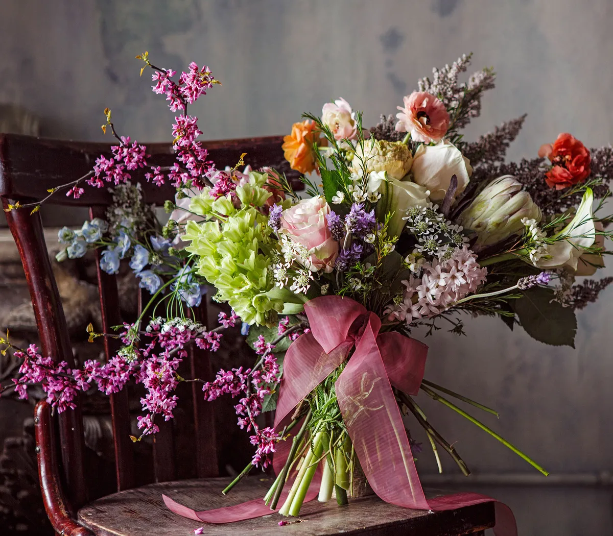 Ein üppiger Blumenstrauß mit Rosen, Grünzeug und zarten Blüten, verziert mit einem rosa Band, steht auf einem Holzstuhl.