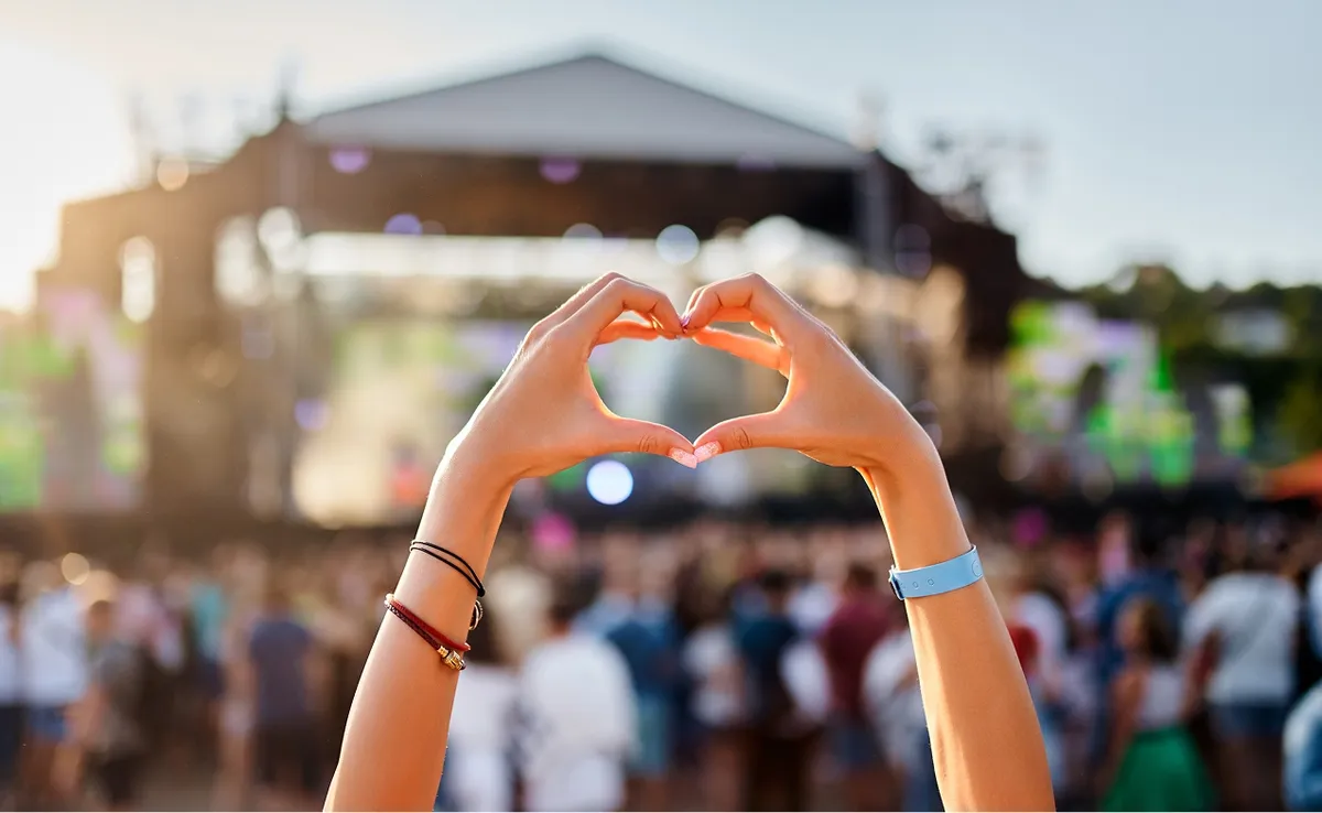 Auf dem Bild zeigt eine Person bei einem Fest mit vielen Besuchern ein Herz aus den Händen, vor einem Bühne-Hintergrund.