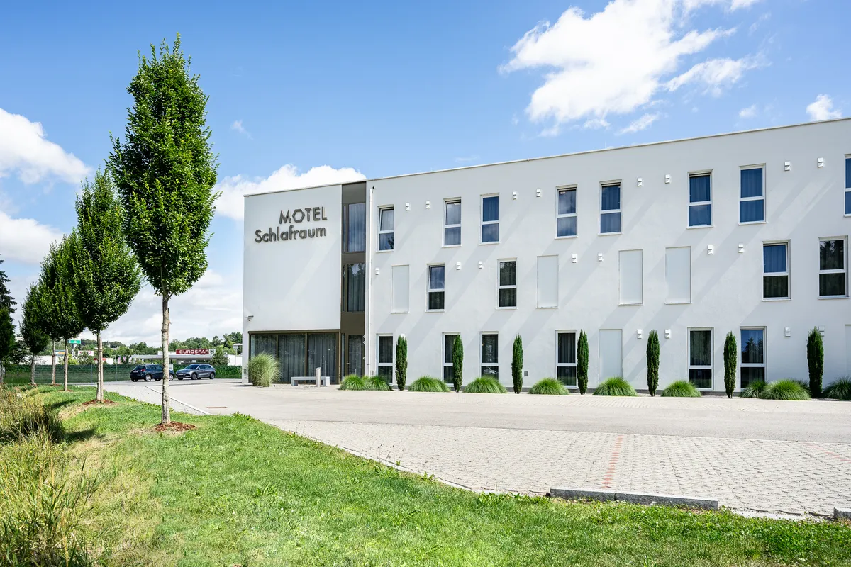 Das Bild zeigt ein modernes Motel mit dem Namen „Motel Schlafraum“ in ruhiger Umgebung mit Grünflächen und Parkplatz.