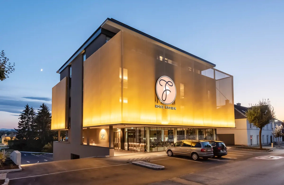 Das Bild zeigt das modern beleuchtete „Hotel Fischer“ mit klarem Design, Glasfront und ruhiger Umgebung bei Abenddämmerung.