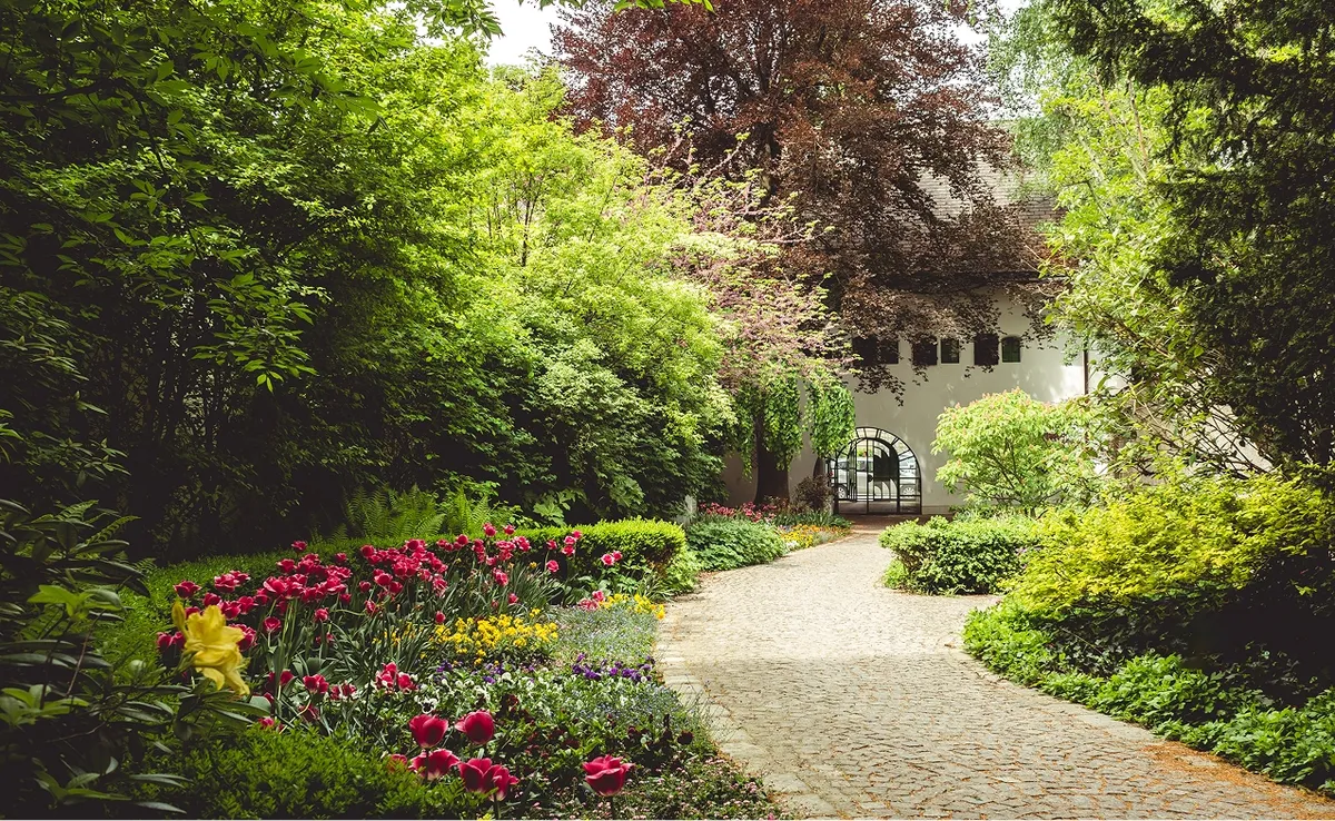 Ein idyllischer Gartenweg führt durch üppige Blumenbeete und grüne Büsche zu einem alten Kornspeicher mit Arkaden.