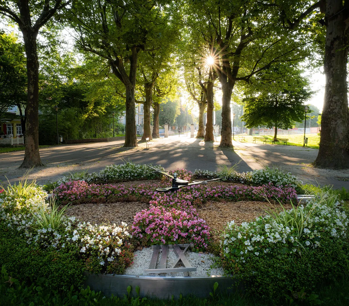 Das Bild zeigt die Blumenuhr im Wiener Volksgarten: Eine kreisförmige Blumenanlage mit Sonnenstand-Anzeige, umgeben von Bäumen und Pfaden.
